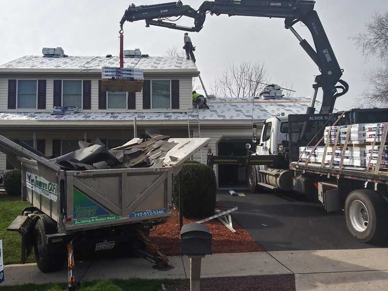 An Equipter and a large truck carrying GAF shingle packs on its back, with a crane that is directly delivering the workers on a roof these shingle packs